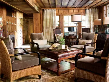 Rustic living room with wood-paneled walls and ceiling, featuring wicker armchairs with plush taupe cushions surrounding a tufted leather ottoman.