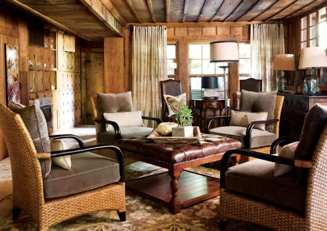 Rustic living room with wood-paneled walls and ceiling, featuring wicker armchairs with plush taupe cushions surrounding a tufted leather ottoman.