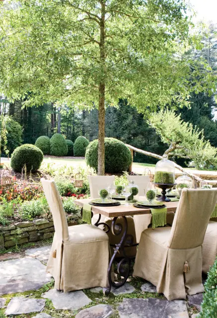 Outdoor dining table set with green-fringed placemats and moss-filled glass cloches, surrounded by manicured boxwood shrubs and lush greenery under a large tree.