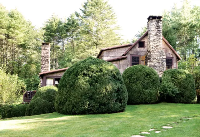 Rustic stone cottage with large stone chimneys, surrounded by manicured green bushes and tall pine trees. A stone pathway leads across the lawn.