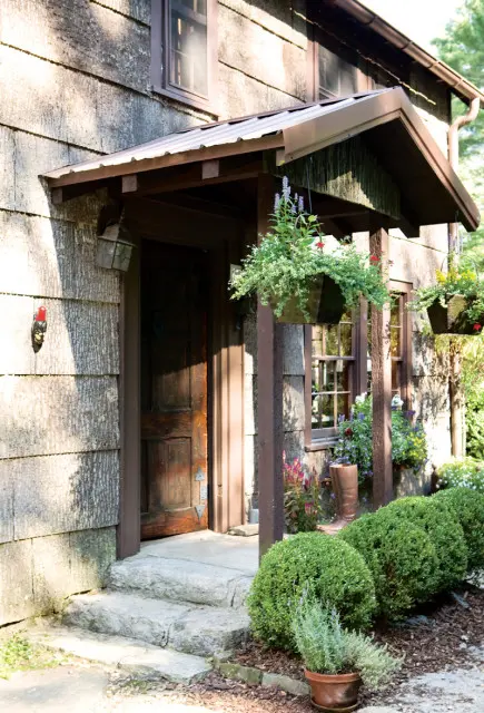 Rustic wooden door with stone steps leading to a house with bark-textured siding and a metal awning adorned with hanging flower baskets.