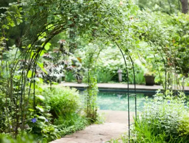 Stone pathway leading through a garden archway covered in greenery, with a swimming pool visible in the background.