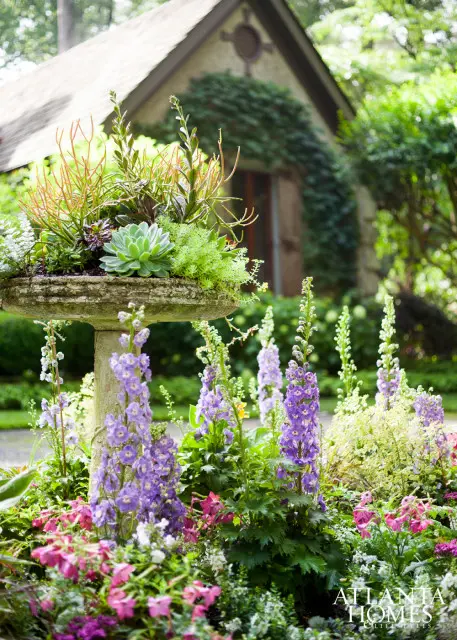 Stone birdbath filled with succulents and ornamental plants, surrounded by purple delphiniums and pink flowers, with a stone cottage in the background.