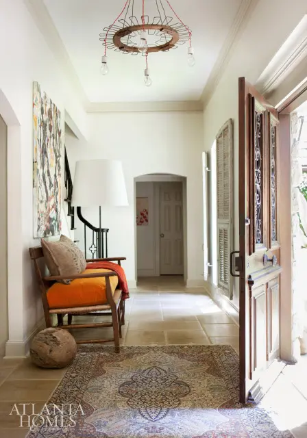 Entryway with stone tile floor, Persian rug, wooden bench with orange cushion and pillows, abstract art, and a unique chandelier.