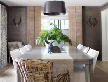 Dining room with a long gray table, white upholstered chairs, and a large planter of ferns topped with a rabbit statue. Textured dark walls feature mounted animal horns.