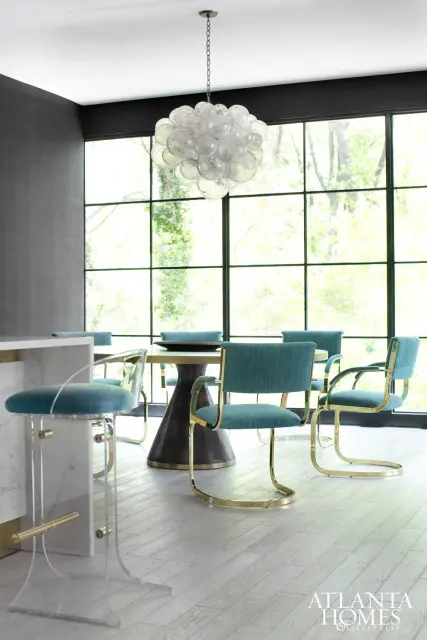 Dining room with teal upholstered chairs with gold frames around a pedestal table, beneath a clear bubble chandelier. Large windows overlook greenery.