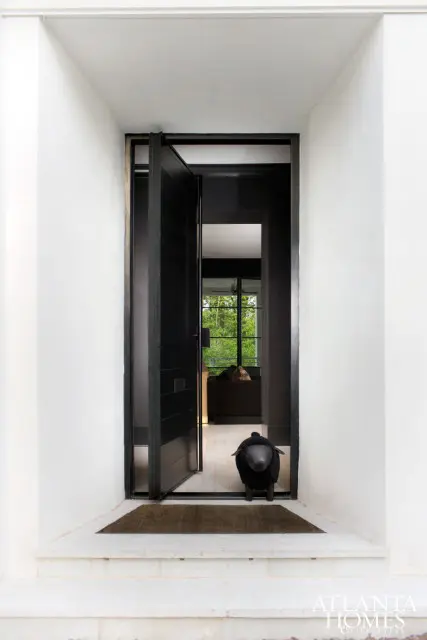 Modern black pivot door opening into a hallway with light wood floors, a black sheep ottoman, and a glimpse of a living room with large windows overlooking trees.