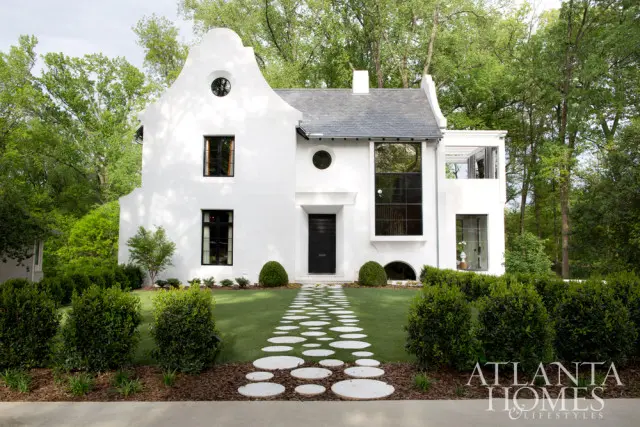 Modern white stucco house with a steep, curved gable roof, black framed windows, and a circular stone pathway leading to the front door. Surrounded by lush green trees and manicured hedges.