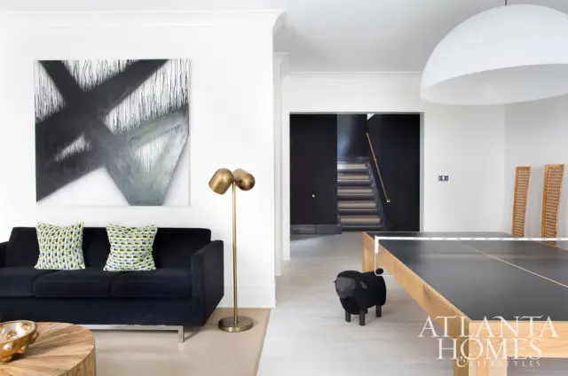Modern living room with black sofa, abstract art, brass floor lamp, ping pong table, and dark wood staircase in background.