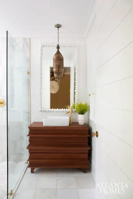 Guest bathroom with a white rectangular vessel sink on a dark wood vanity with ornate detailing. A brass faucet and a decorative Moroccan lantern hang above. Marble tile floors and a glass shower enclosure are visible.