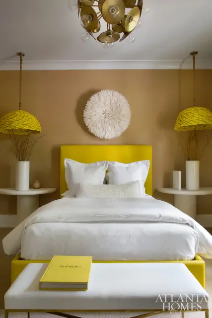 Guest bedroom with a bright yellow upholstered bed, white bedding, and a white bench. The walls are a warm tan, and yellow woven pendant lights hang on either side of the bed.