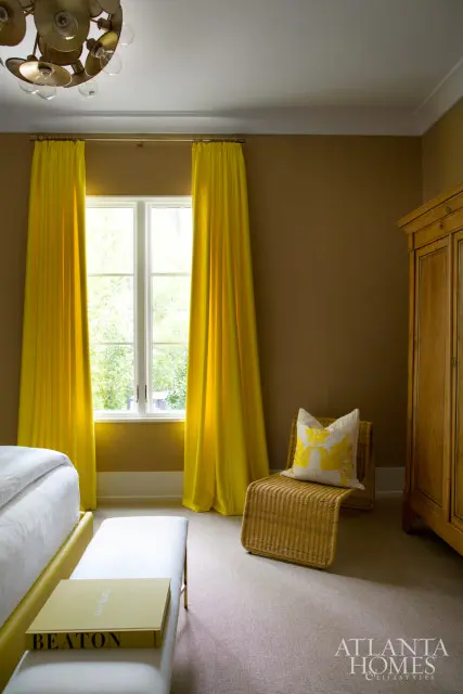Guest bedroom with brown walls, bright yellow curtains, a wicker chair with a floral pillow, a wooden armoire, and a white bench with a book.