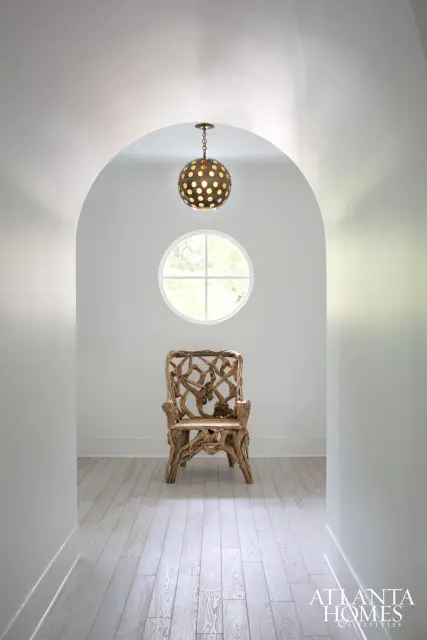 Hallway with white walls, light wood floors, a round window, a globe pendant light, and a chair made of natural wood branches.