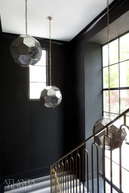 Dark hallway with geometric pendant lights and brass railing on staircase. Large windows overlook greenery.