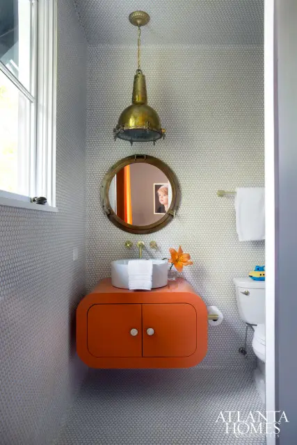 Powder room with white penny tile walls and floor, featuring a floating orange vanity with a white vessel sink, brass fixtures, and a nautical porthole mirror with a brass frame.