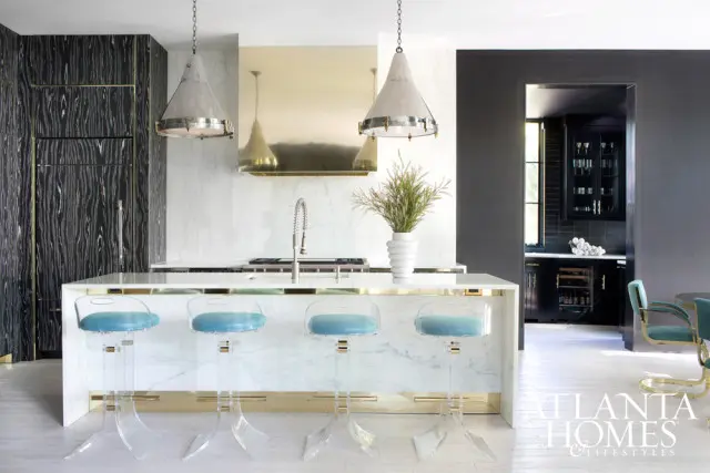 Modern kitchen with a white marble island, clear acrylic bar stools with blue upholstery, and dark patterned cabinetry. A brass range hood is mounted above the cooktop.