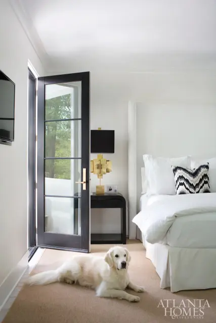White bedroom with a large bed, black french doors open to greenery, a golden retriever lies on the tan carpet, and a gold geometric lamp sits on a black nightstand.