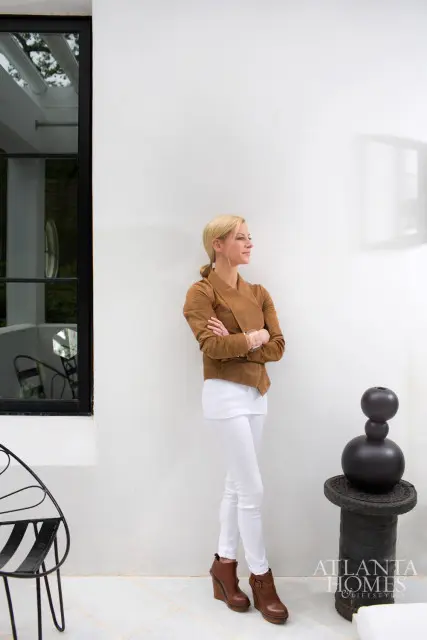 Woman with blonde hair in a ponytail wearing a tan suede jacket and white pants, standing with arms crossed next to a white wall.