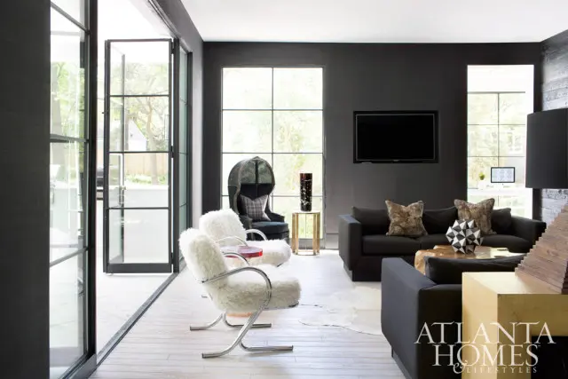 Dark gray living room with black sofas, white boucle armchairs, and large grid windows. A dark green armchair with a plaid pillow sits near a gold side table.