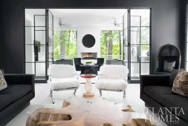Modern living room with black sofas, white fluffy armchairs, a large wood slab coffee table, and glass-paneled doors opening to a sunlit patio with a fireplace.