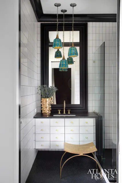 Bathroom with white subway tile walls, black hex tile floor, and a vanity with silver drawers and brass hardware. A gold vase with lavender sits on the counter, and a cluster of blue glass pendant lights hang above the mirror.