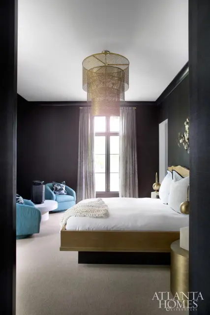 Bedroom with dark walls, a gold platform bed, white bedding, and a large tiered gold chandelier. Two teal armchairs sit near a window with patterned drapes.