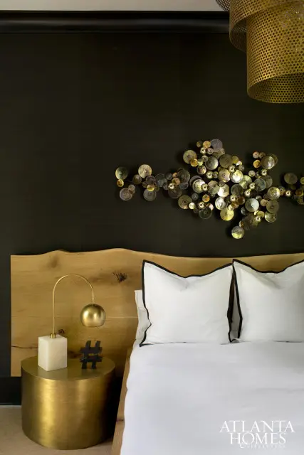 Bedroom with a live-edge wood headboard, dark textured wallpaper, and a metallic wall sculpture. A brass bedside table holds a lamp and a hashtag decor piece.