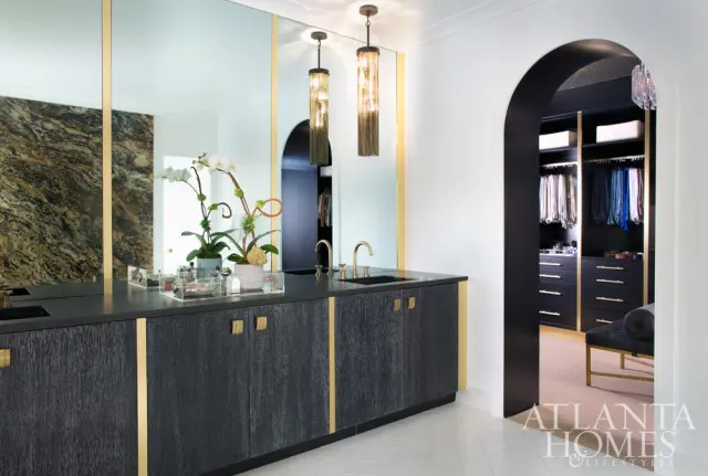 Master bathroom vanity with dark wood cabinets, brass hardware, and black countertop. A walk-in closet with dark cabinetry and organized clothing is visible through an arched doorway.