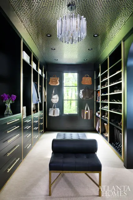 Black master closet with gold-trimmed shelves, drawers, and a geometric gold-patterned ceiling. Features a crystal chandelier, black bench, and handbags displayed on the wall.