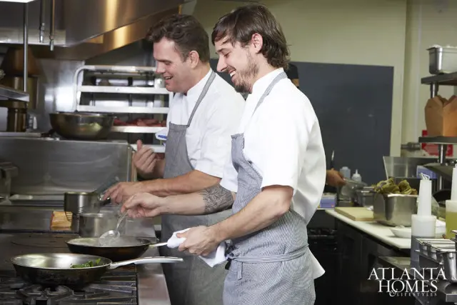 Two chefs in a commercial kitchen wearing aprons and chef coats, laughing while cooking. Stainless steel surfaces and equipment are visible.