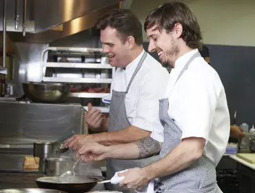 Two chefs in white coats and aprons work in a commercial kitchen, one stirring a pan and the other holding a towel.