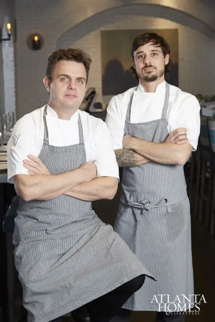 Two chefs in white chef coats and striped aprons stand with arms crossed in a restaurant dining room.