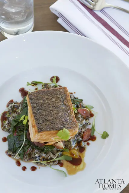 Crispy skin salmon fillet served over wild rice and greens, drizzled with sauce, on a white plate. Atlanta Homes & Lifestyles logo in the bottom right corner.