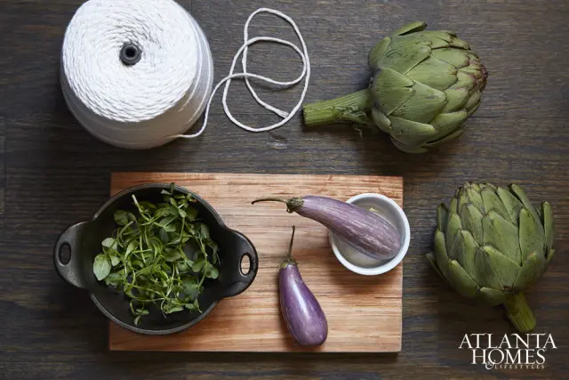 Two purple eggplants and two artichokes arranged on a wooden surface with a spool of white twine and a bowl of microgreens.