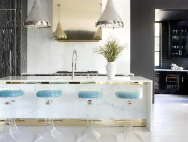 Modern kitchen with white marble island, clear acrylic bar stools with blue cushions, and a black wood-grain refrigerator.