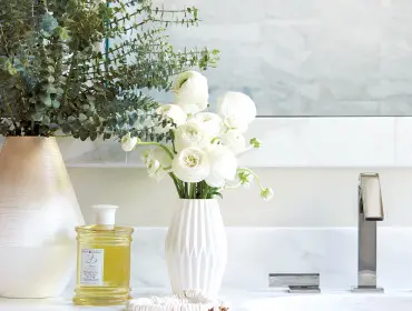 White marble bathroom vanity with a brushed nickel faucet, a white faceted vase filled with white ranunculus flowers, a large vase with eucalyptus, a bottle of Royal Apothecary bath oil, and a shell necklace.