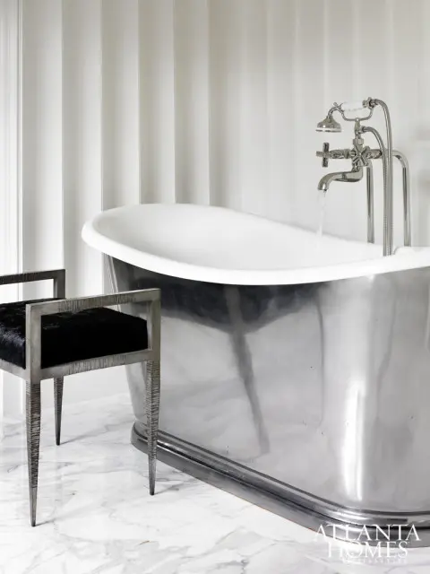 Modern bathroom with a freestanding polished metal bathtub, a black upholstered bench with a metal frame, and striped wallpaper. Water flows from a chrome faucet into the tub.