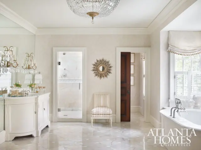 Elegant bathroom with a white vanity, crystal chandelier, glass-enclosed shower, and a freestanding bathtub. The floor features marble tiles in a geometric pattern.