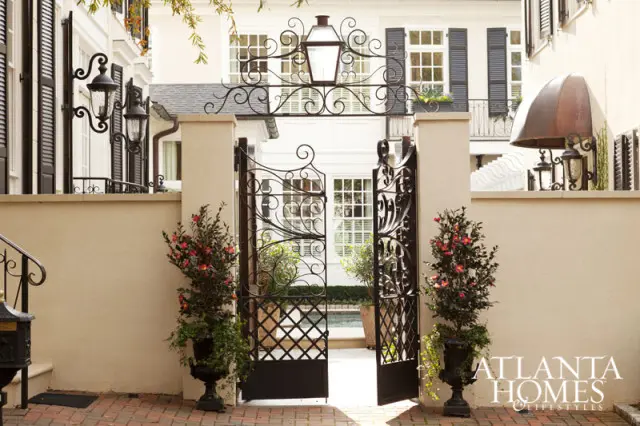 Wrought iron gate with ornate scrollwork and a lantern above, flanked by potted flowering bushes and urns.