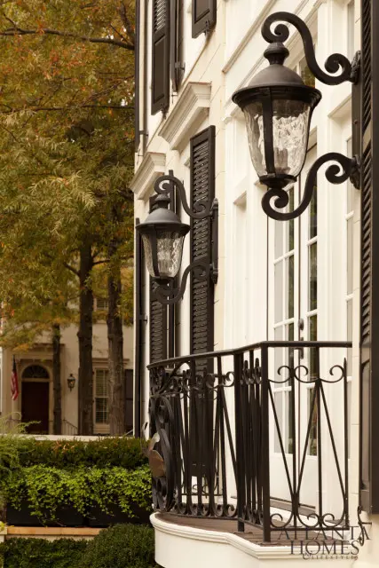 White house exterior with black shutters, ornate wrought iron balcony, and decorative sconce lighting. Lush green landscaping in foreground.