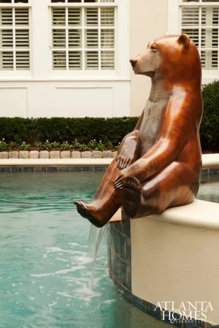 Bronze bear sculpture with water flowing from its feet into a swimming pool, with a building and manicured bushes in the background.