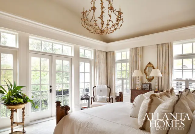 Bedroom with white bedding, ornate gold chandelier, French doors opening to a balcony, and patterned curtains.