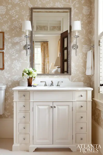Cream vanity with double doors and multiple drawers, topped with a white countertop and double sinks. Floral wallpaper and sconce lighting adorn the walls.