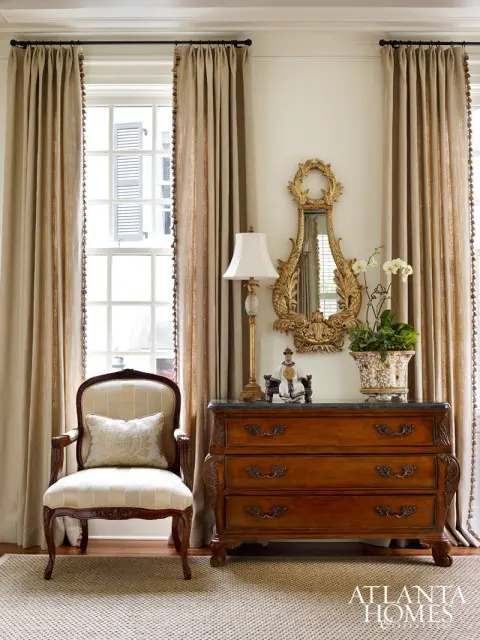 Traditional living room with a cream upholstered armchair, a wooden chest of drawers with a marble top, and ornate gold mirror above.