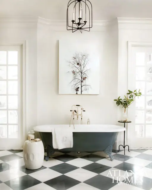 White bathroom with a dark gray clawfoot tub, black cage chandelier, and black and white checkered marble floor. Artwork of a bare tree hangs above the tub.