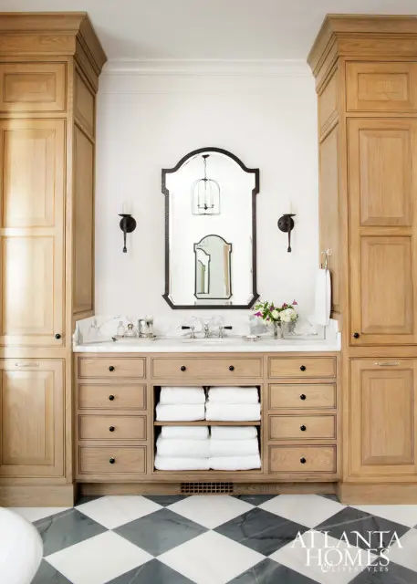 Light wood vanity with marble countertop, black hardware, and built-in shelving for towels. Flanked by tall wood cabinets. Above, an ornate mirror reflects a birdcage chandelier.