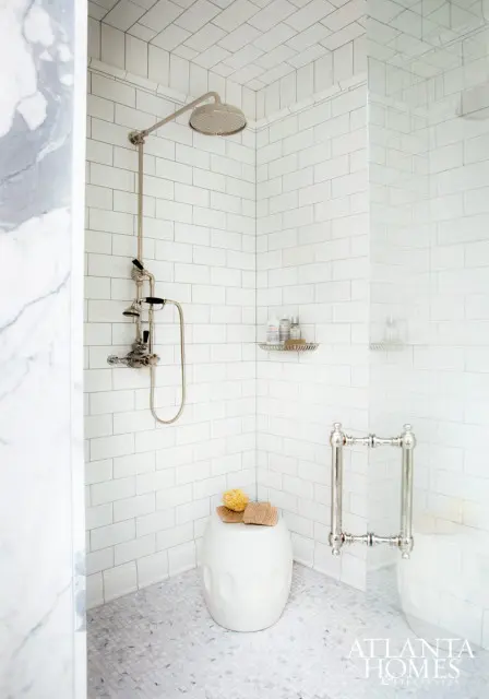 White subway tile shower with chrome fixtures, a glass door, a white stool with sponges, and marble mosaic tile floor.