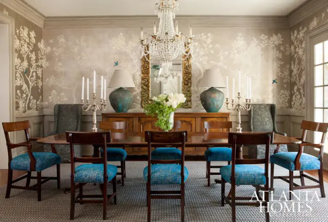 Dining room with a large wooden table and chairs upholstered in blue fabric, set against a wall with a tree and bird motif wallpaper. A crystal chandelier hangs above.