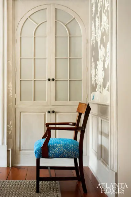 Wooden armchair with bright blue patterned upholstery in front of a tall, arched cabinet with glass-paned doors and a wall with floral wallpaper.