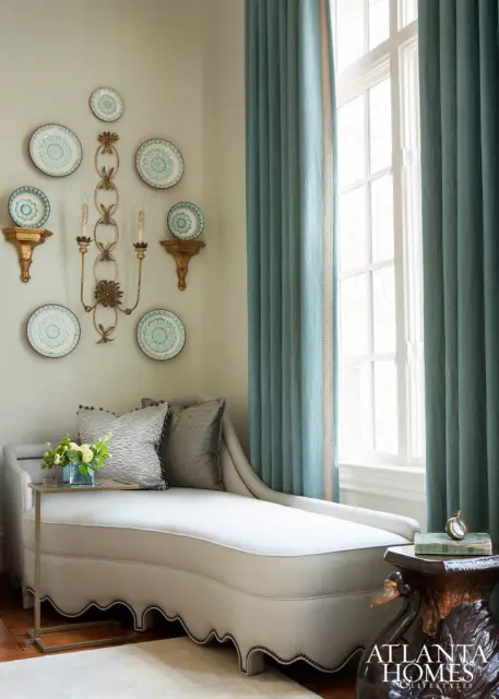 Light gray chaise lounge with decorative trim, flanked by teal curtains and a wall adorned with decorative plates and a sconce.